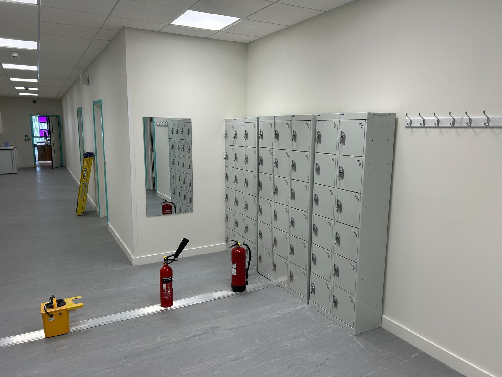 Hallway with gray lockers, red fire extinguishers, and a yellow caution sign on a tiled floor
