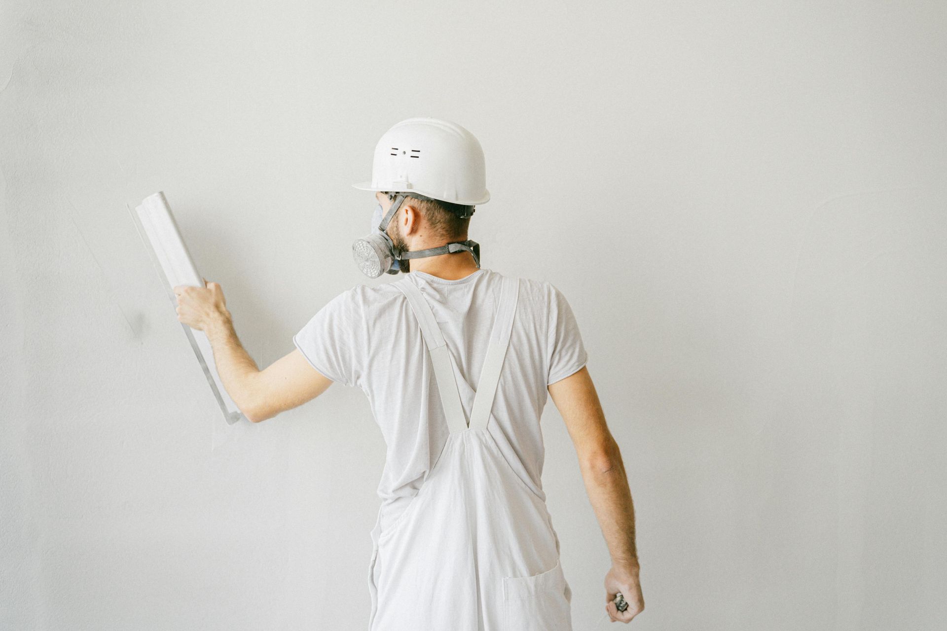 A worker wearing white overalls and a hard hat uses a tool to smooth a white wall.