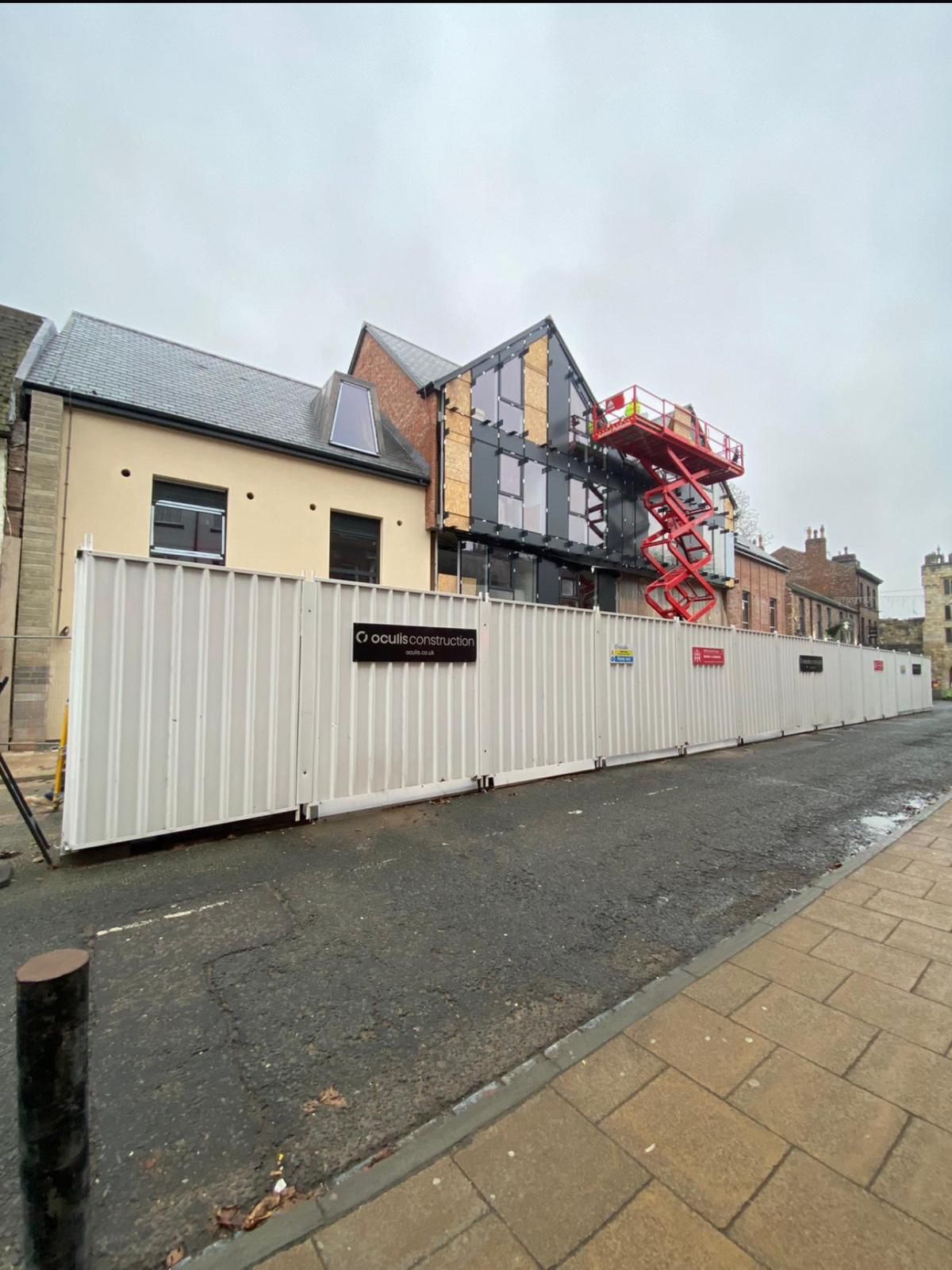 White construction hoarding along a wet street beside old buildings and scaffolding under an overcast sky