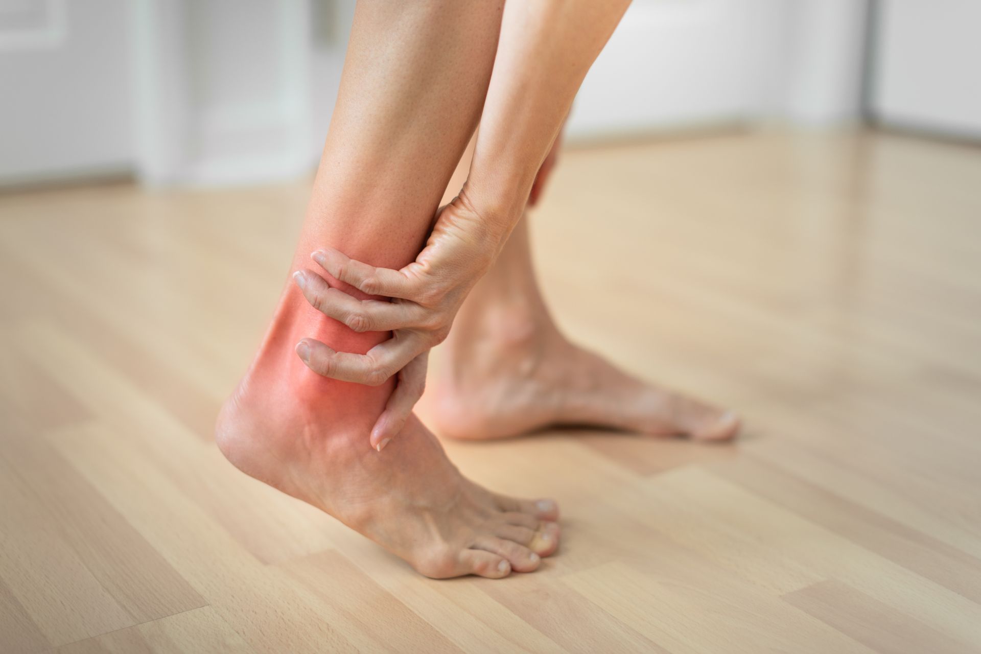 A person's hands holding their ankle, which is highlighted in red to indicate pain or inflammation.