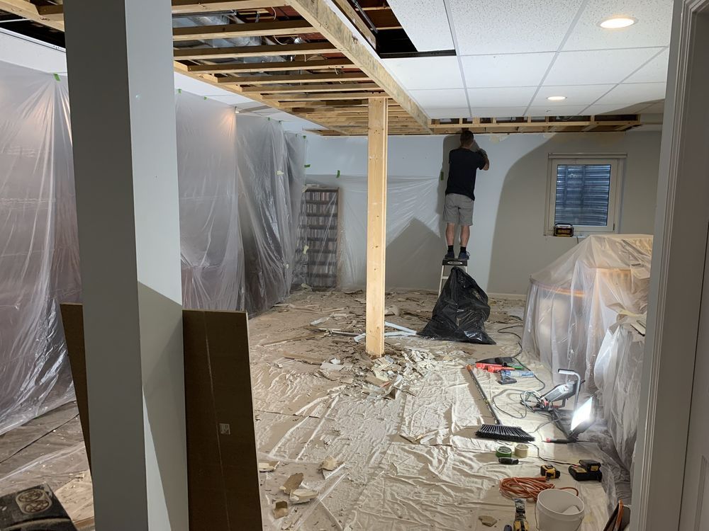 A person on a ladder is working on a partially demolished ceiling in a basement.
