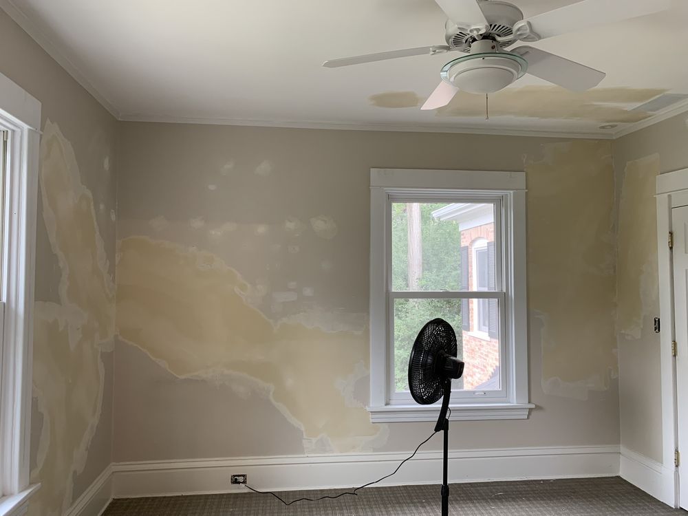 Room with water damage on walls, ceiling, and baseboards; a fan is blowing.