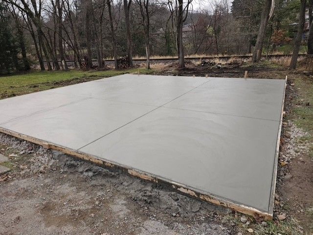 A freshly poured, gray concrete slab for a structure, surrounded by wooden framing and a dirt yard.
