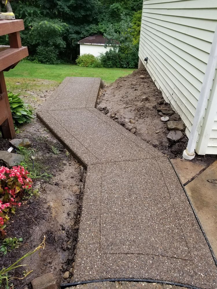 Concrete walkway next to a light yellow house and brown deck, with exposed dirt and grass.