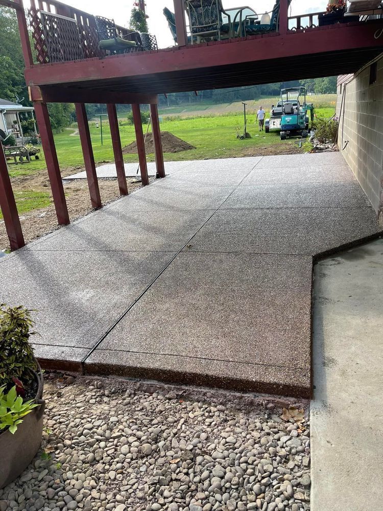 Concrete patio under a deck. Gravel edging, brown deck supports, and a grassy background.