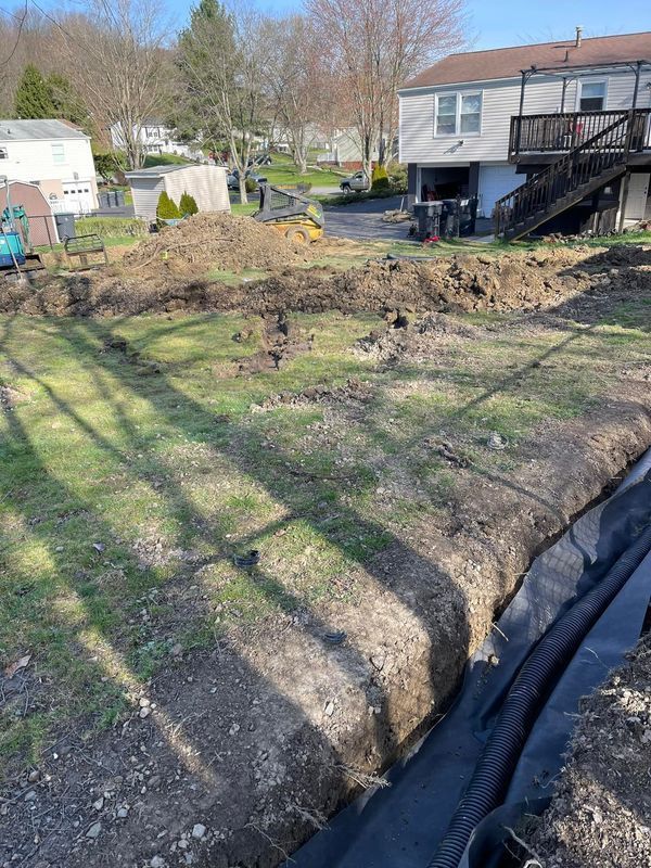 Backyard excavation with trenches, black fabric, and exposed dirt. Houses and a sunny sky in the background.
