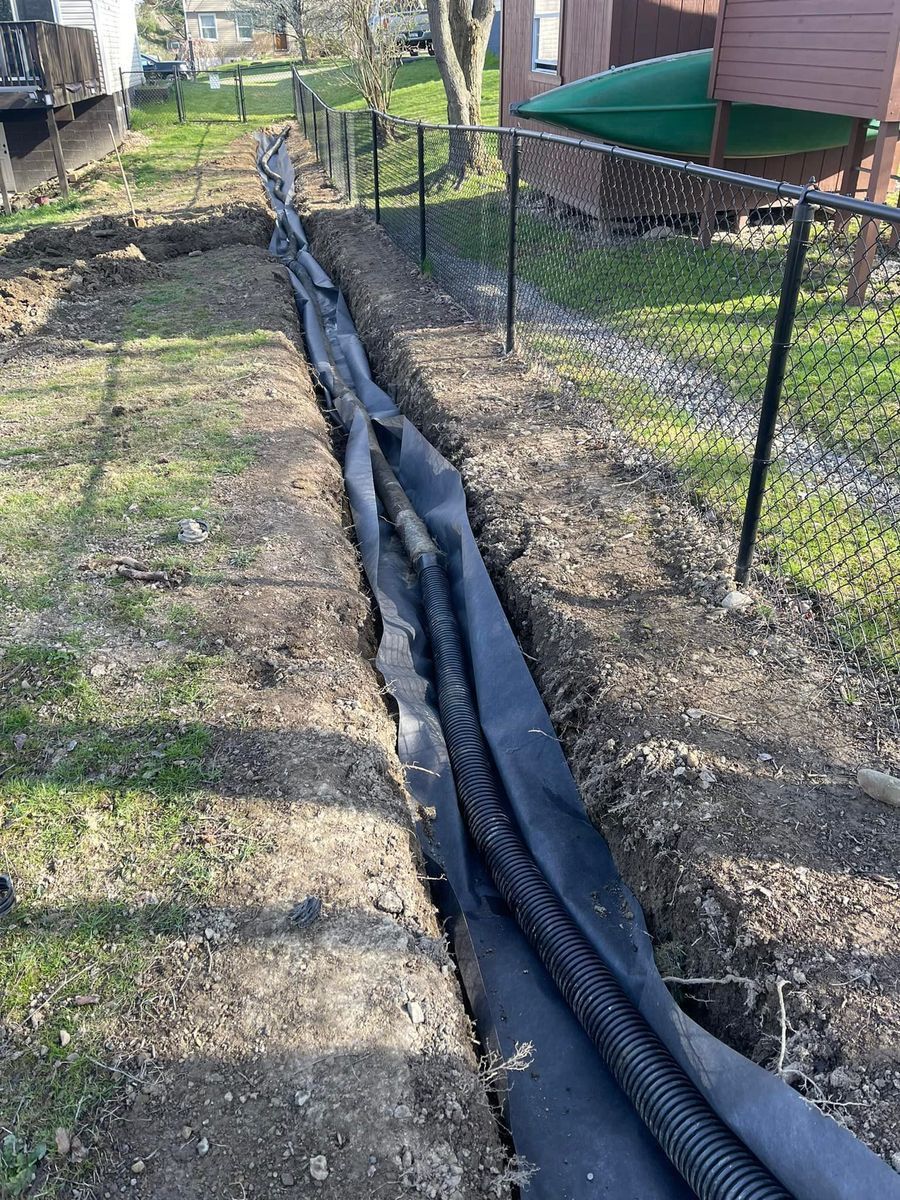 Trench in a yard, containing a black corrugated drainage pipe wrapped in black fabric, next to a black fence.