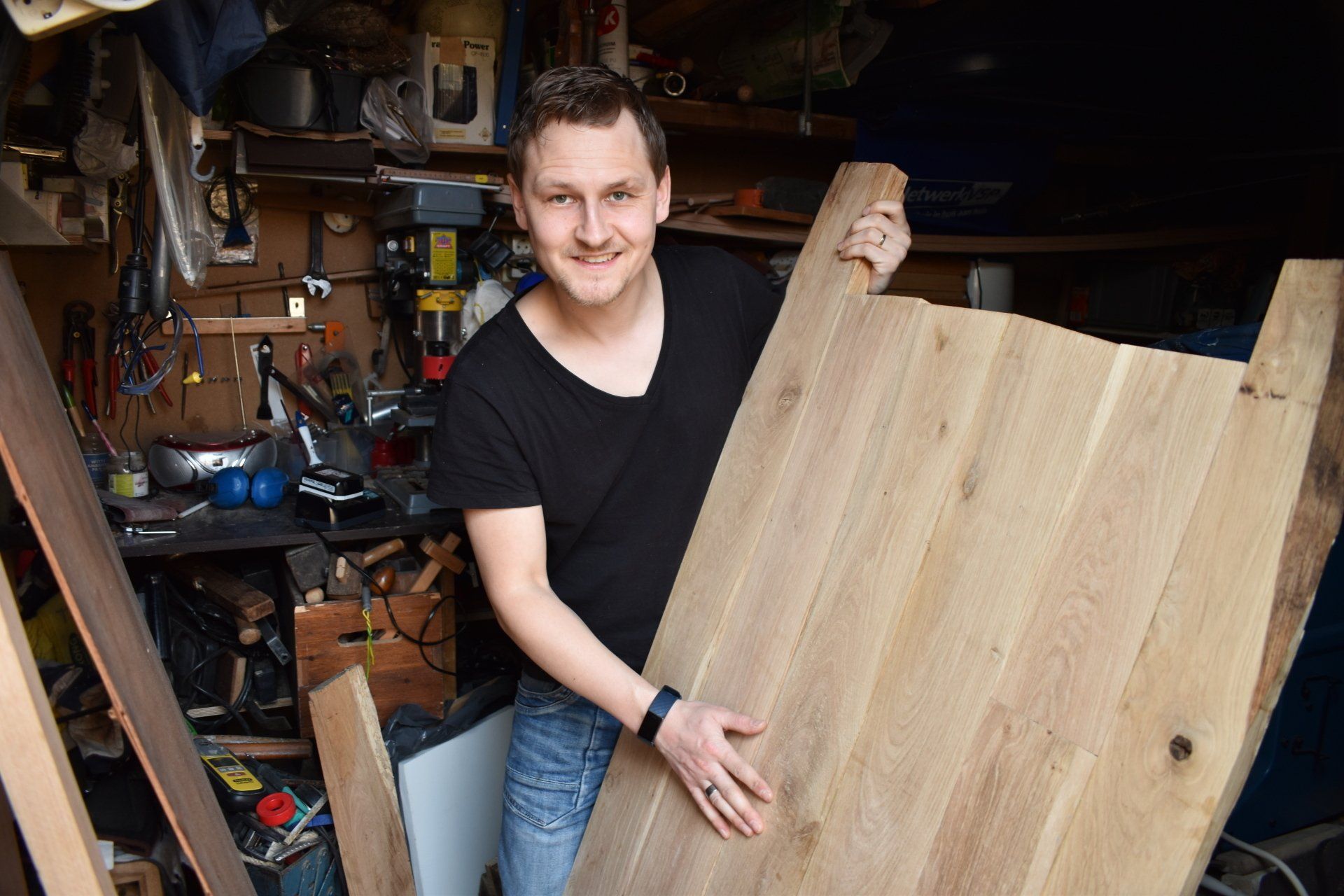 Man in a workshop holding a large wooden slab, smiling. Tools and equipment visible in the background.