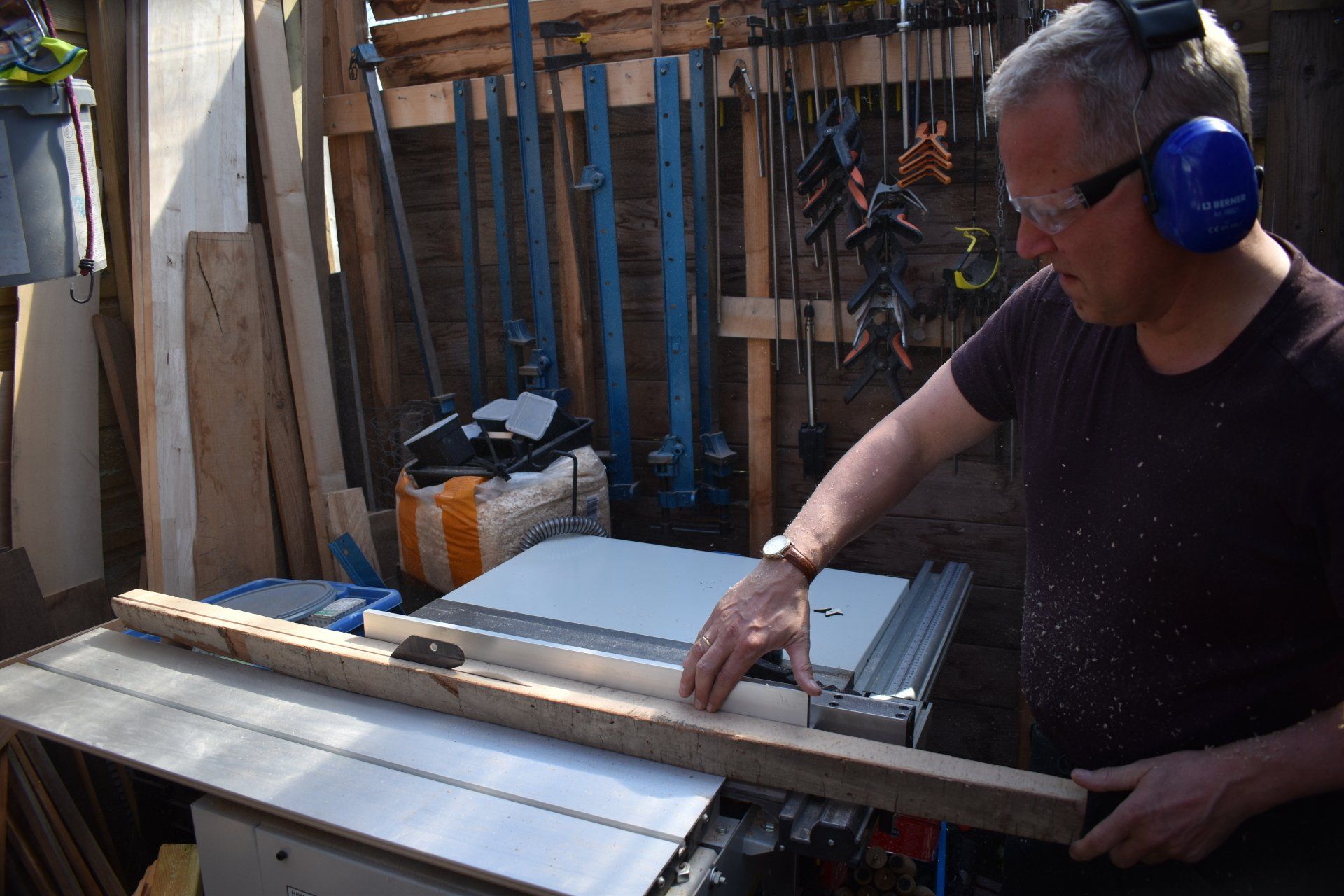 Man using a table saw to cut wood in a workshop, wearing safety glasses and earmuffs.