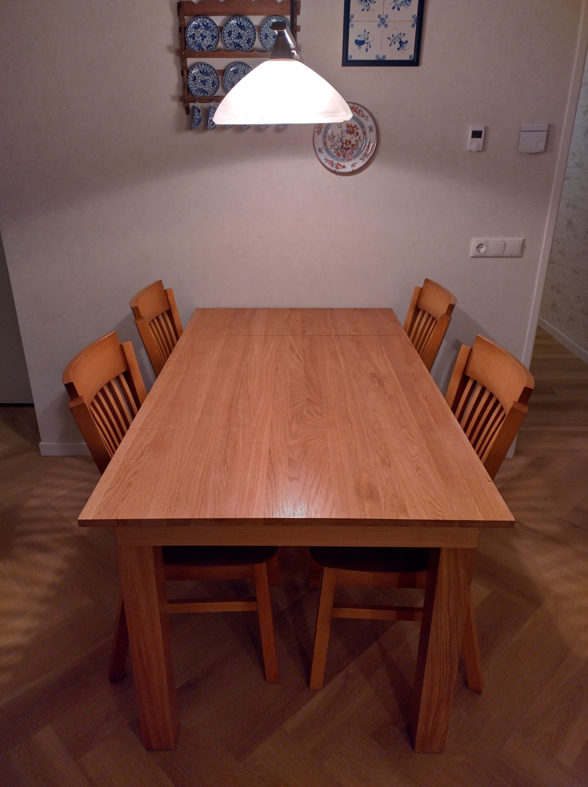 Wooden dining table with four chairs, set in a room with light-colored walls and wood flooring.