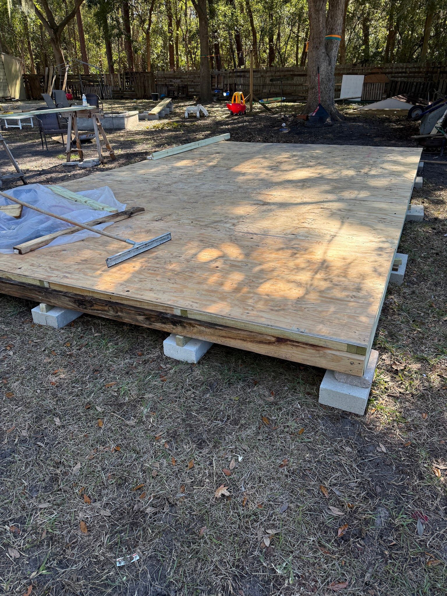 Wooden platform foundation on gravel in a wooded yard, with stacked materials on one corner.