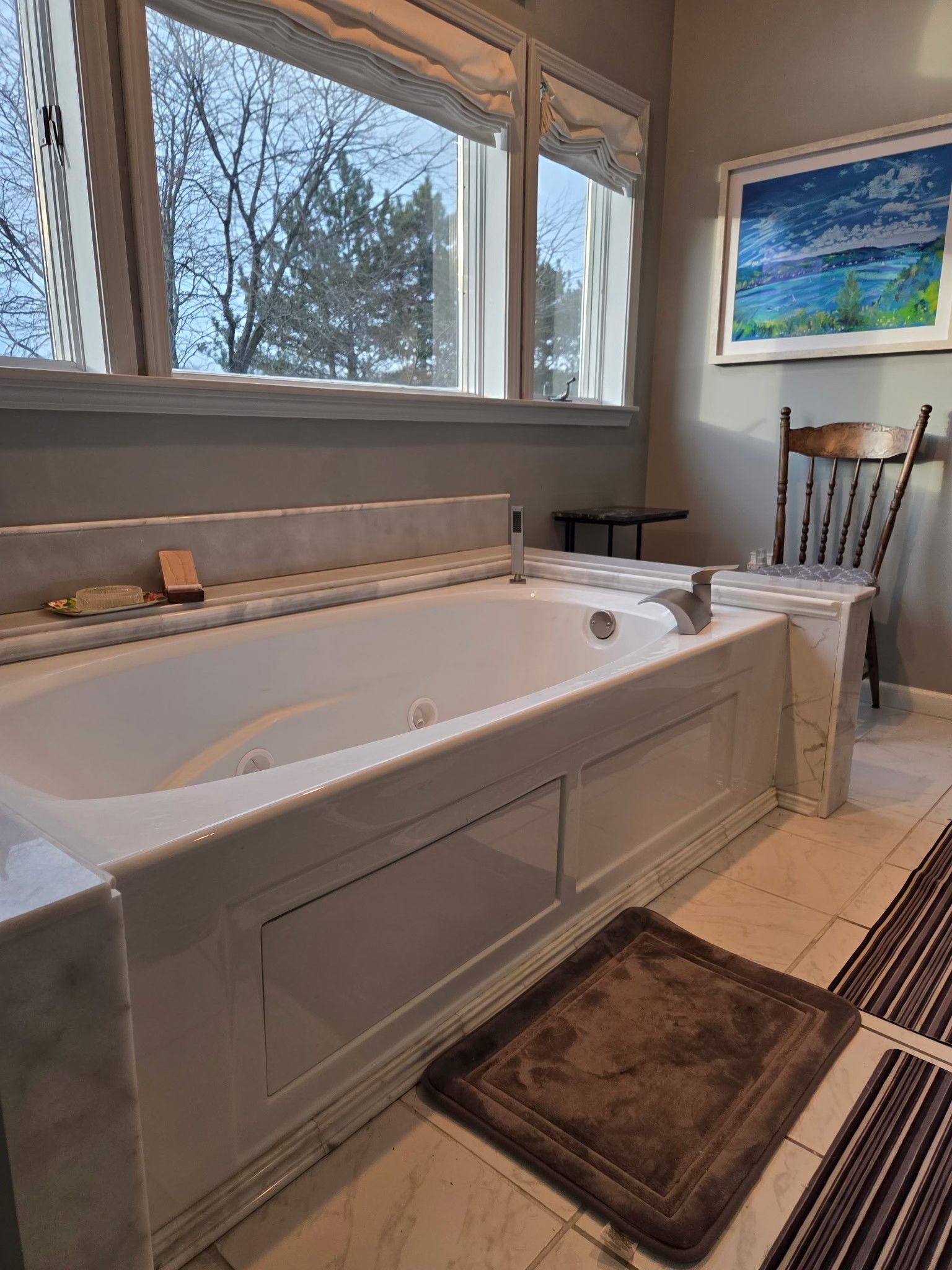 Bathroom with a white soaking tub by a window, bath mat on the floor, and a chair beside it