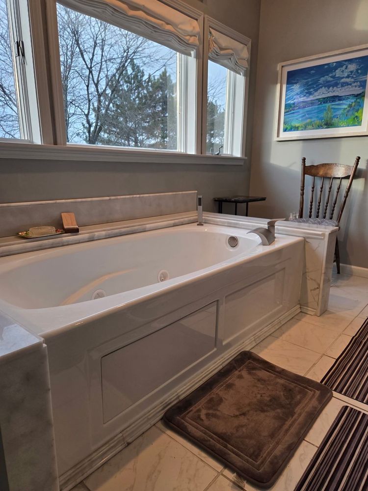 Bright bathroom with a large soaking tub beside windows, a wooden chair, and a landscape painting.