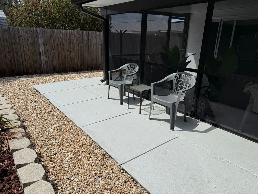 Patio with two gray chairs beside a glass-walled house, bordered by gravel and a wooden fence.