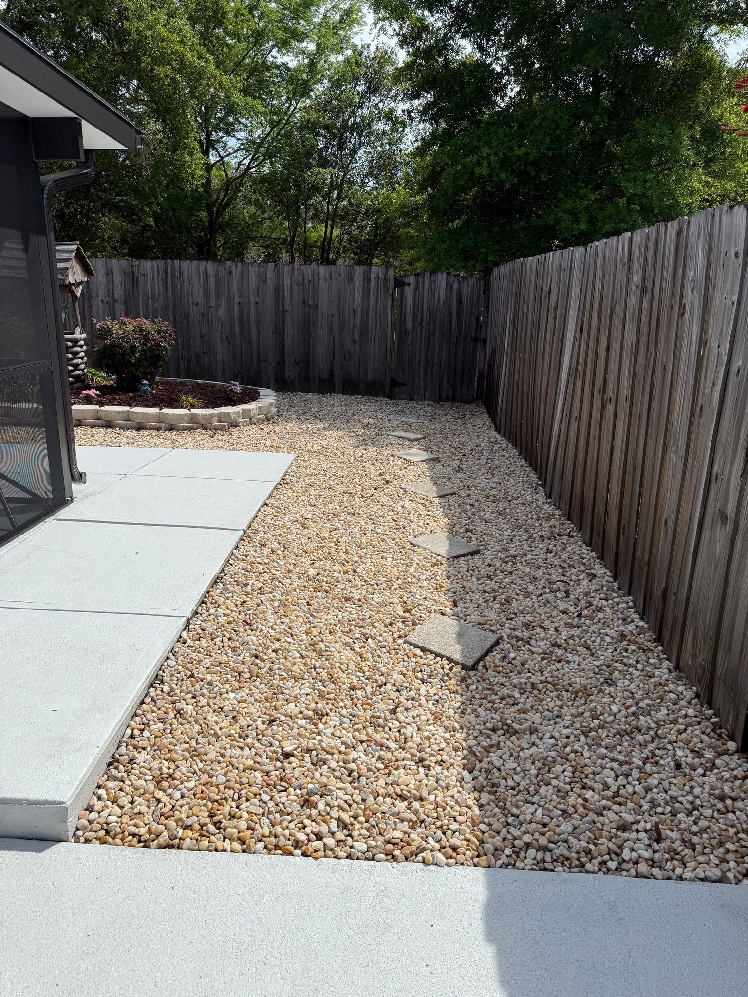 Narrow side yard with gravel path, concrete patio edge, and wooden privacy fence under trees