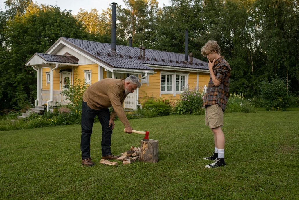 Two people by a small campfire in a grassy yard beside a yellow house, one tending the fire with an axe.