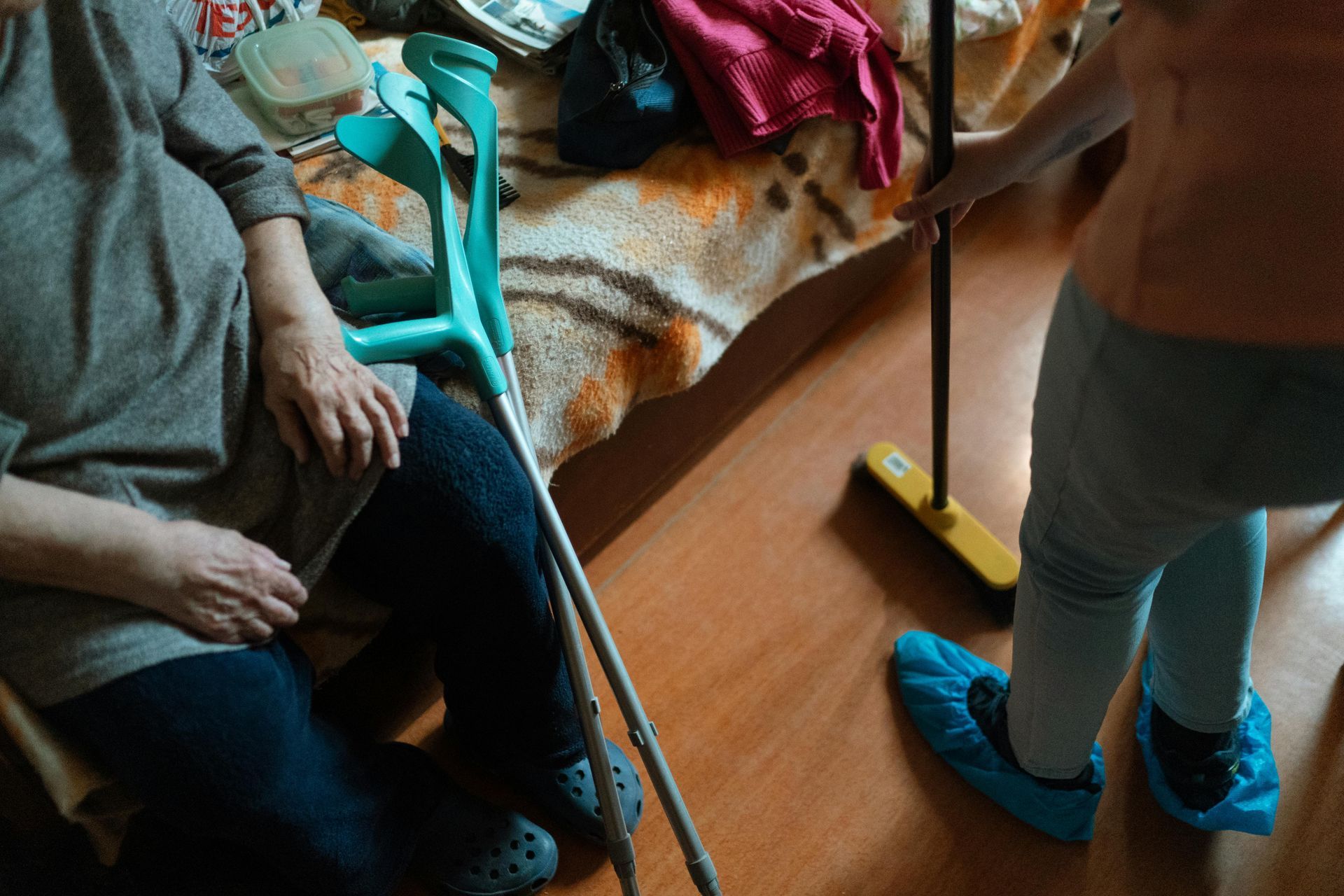 Two people standing and sitting in a room with a baby walker, toys, and a broom on a wooden floor