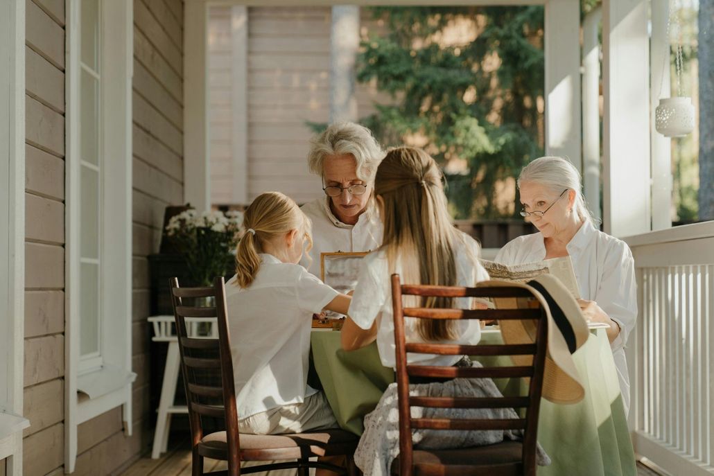 Four people sit on a porch around a table, reading and talking in warm sunlight.