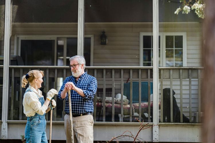 Two people talking on a porch, holding drinks, with chairs and a railing behind them.