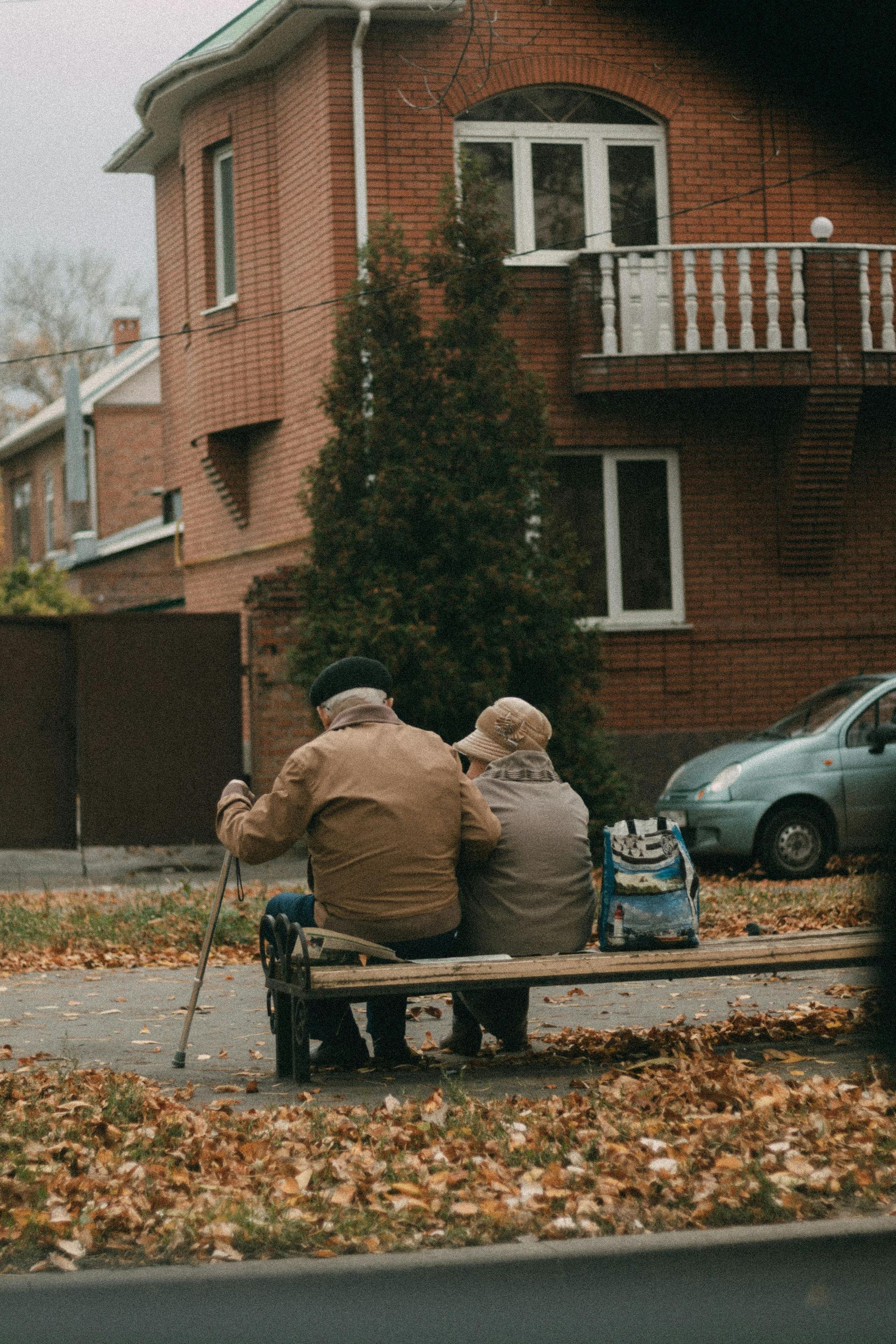 Two people sit on a bench in front of a brick apartment building with fallen autumn leaves.