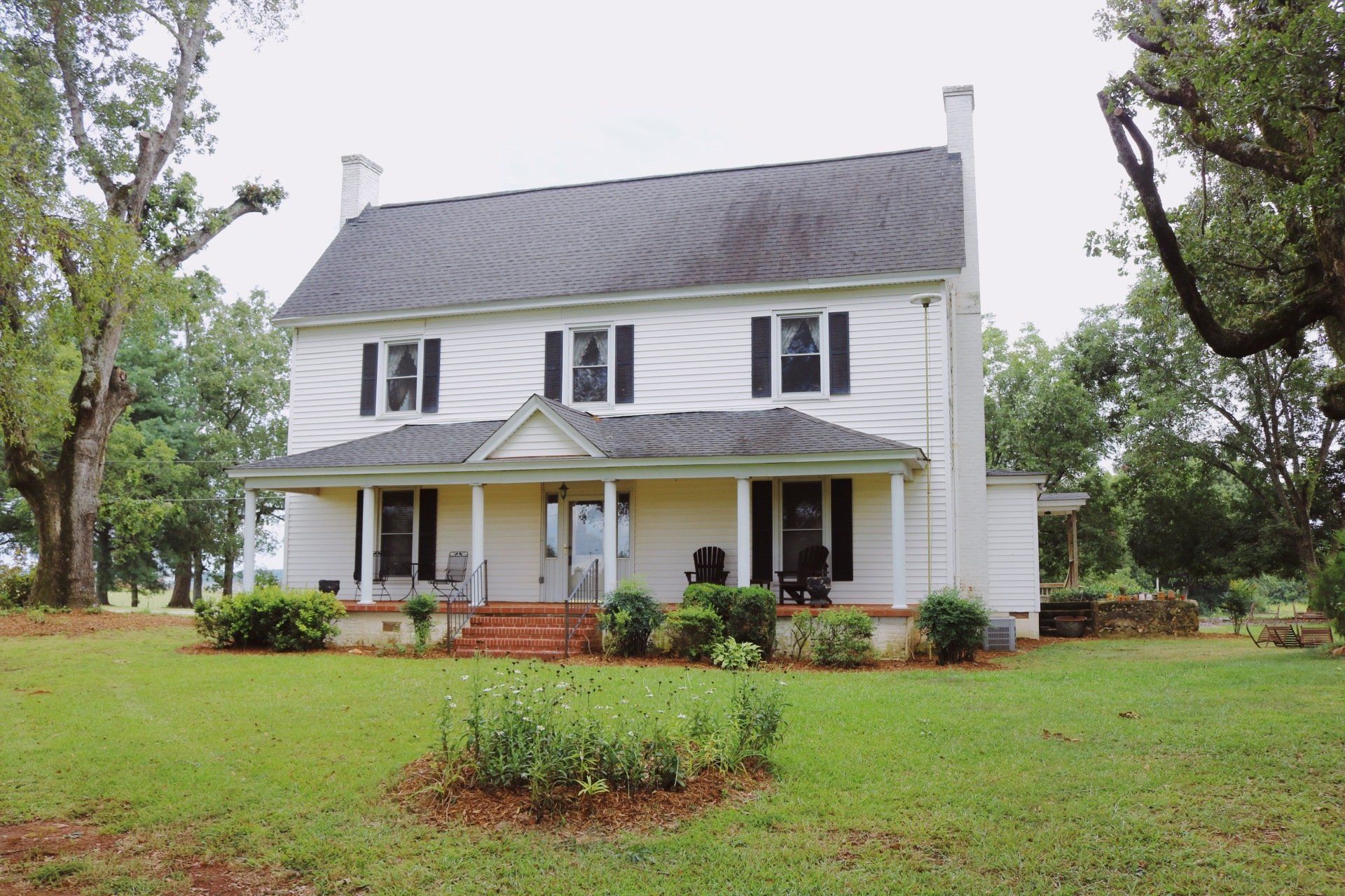 White two-story house with a porch and black shutters, set on a green lawn with trees in the background.