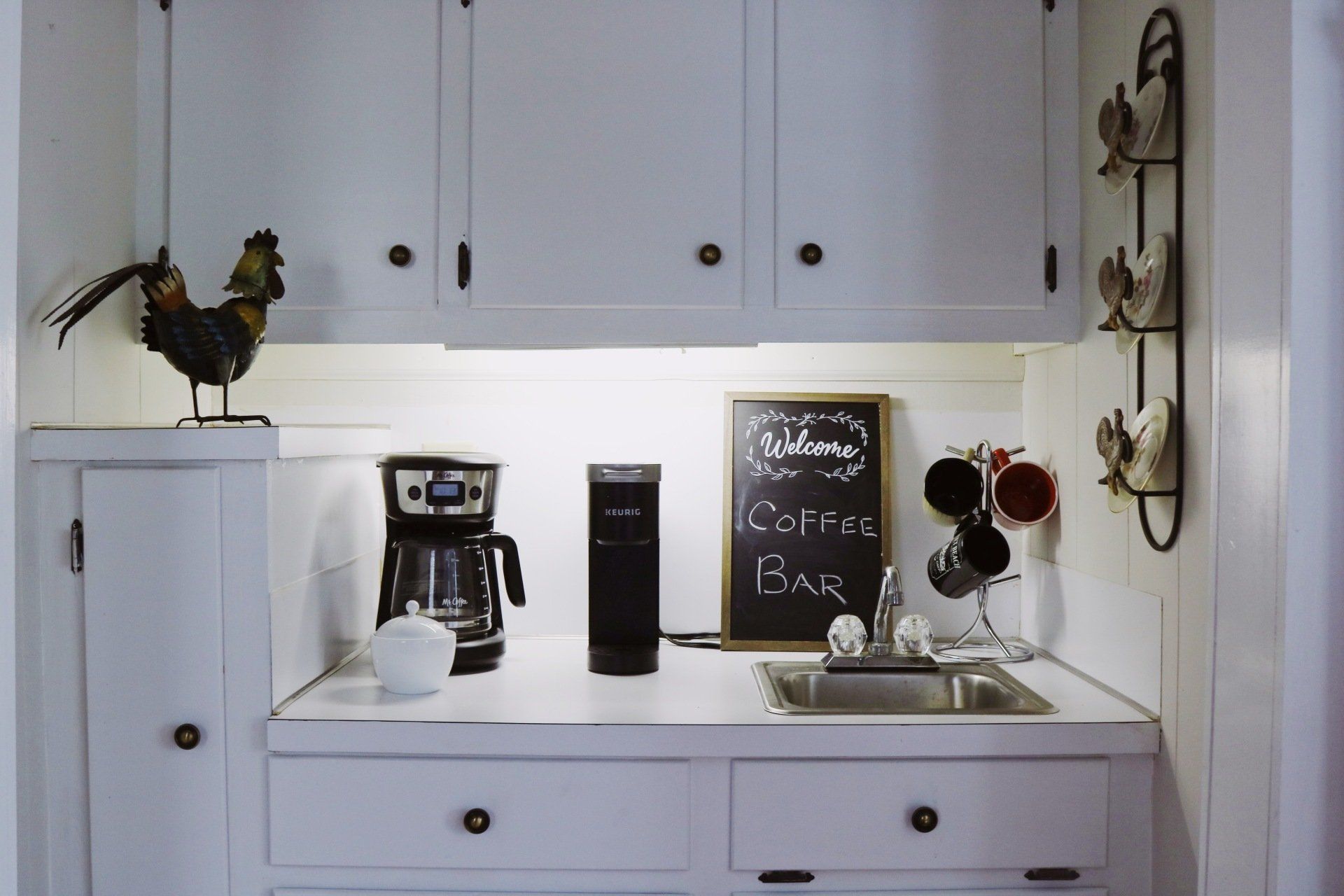 White coffee bar with cabinets, a chalkboard sign, and a rooster statue.