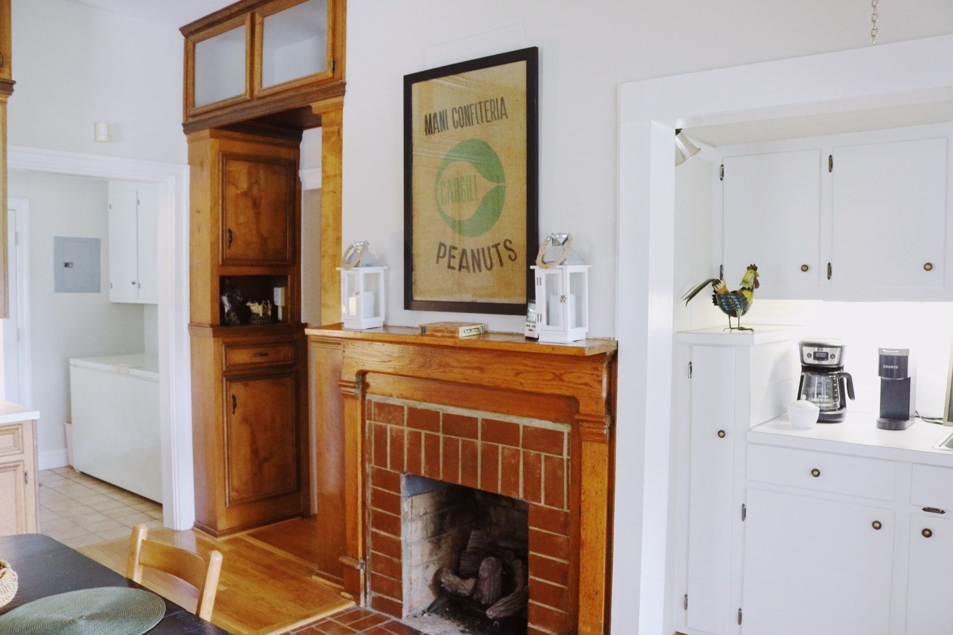 Fireplace with brick, wooden mantel, and a vintage peanut ad, next to a white kitchen.