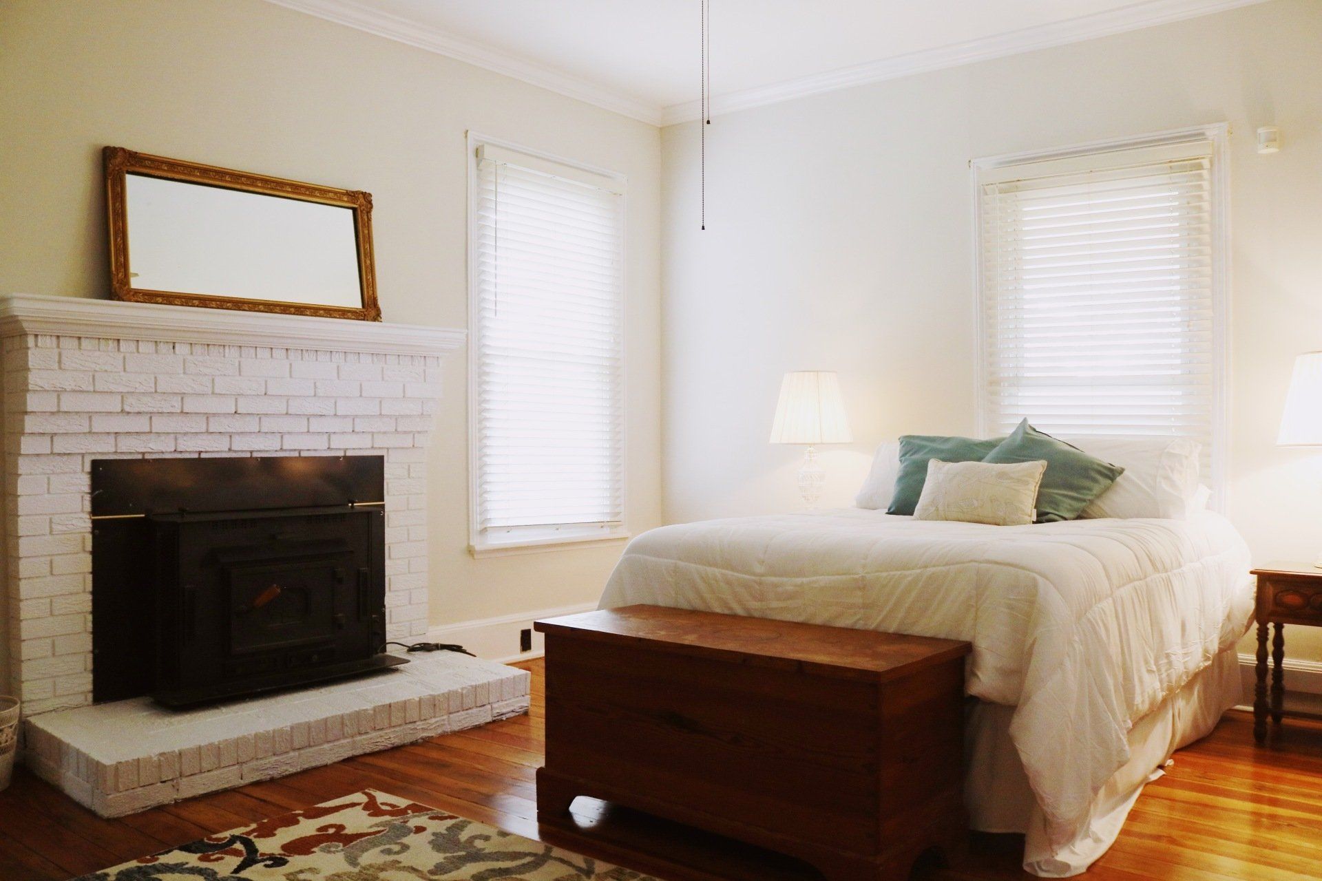 Bedroom with fireplace, bed, and wooden chest. White walls, wood floor, and two windows with blinds.