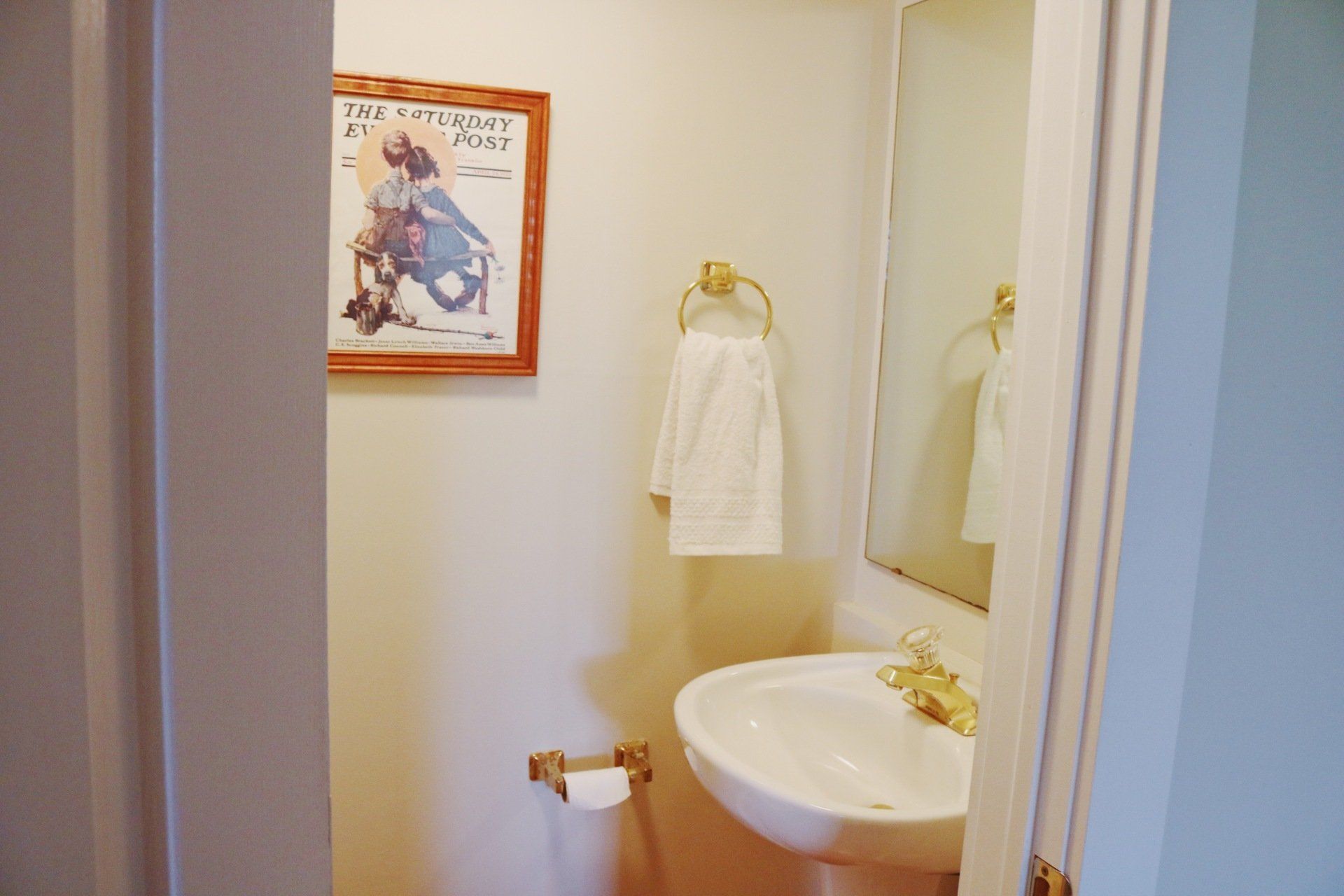 Small bathroom with sink, mirror, and artwork; white walls, gold fixtures.