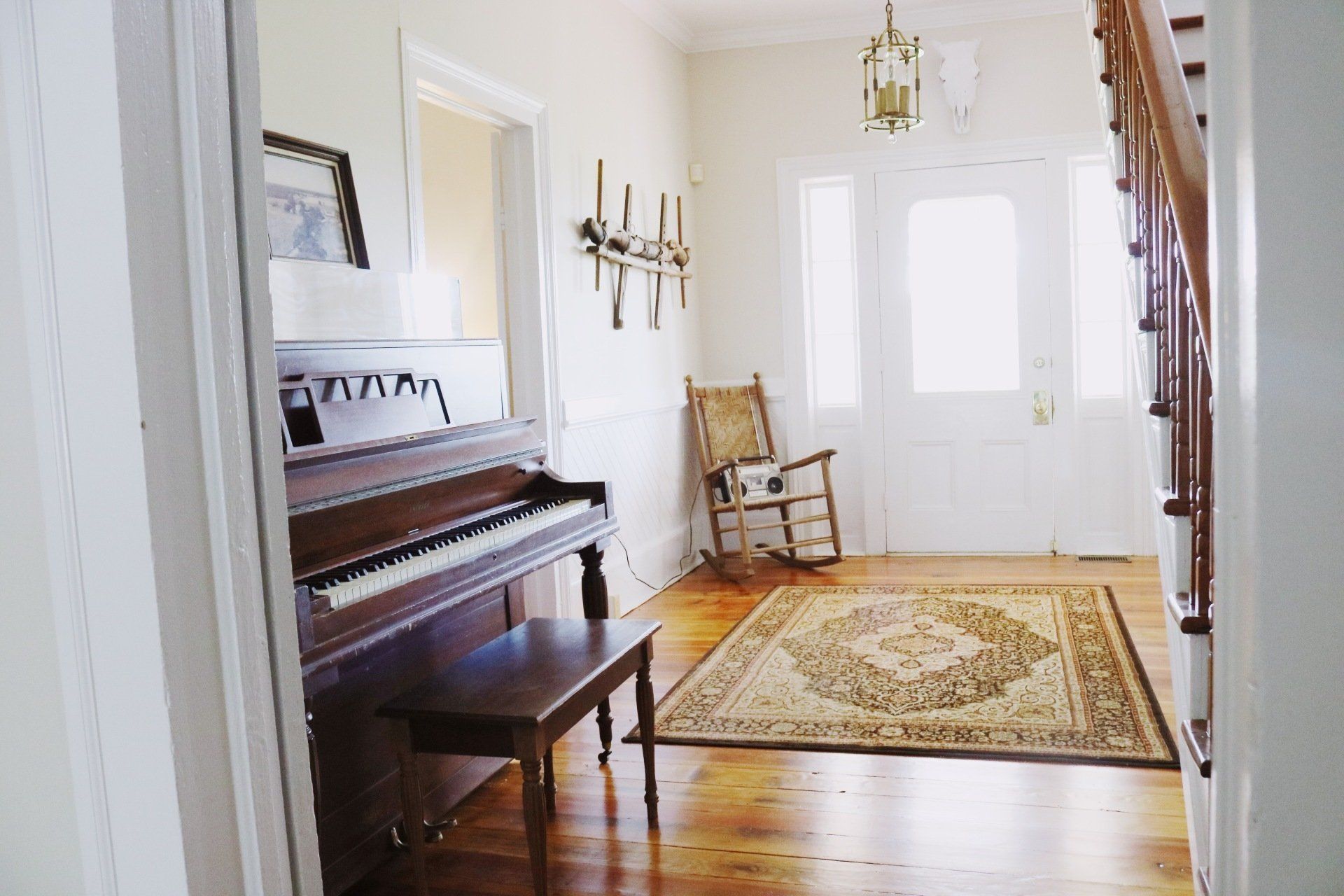 Entryway with piano, rug, and rocking chair. Wooden floors, white walls, and bright door.