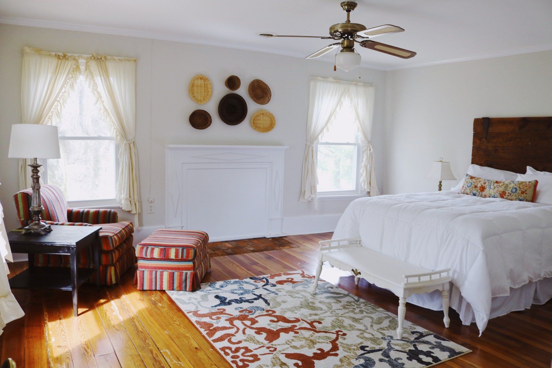 Spacious bedroom with white walls, wood floors, bed, fireplace, windows, and decorative wall baskets.