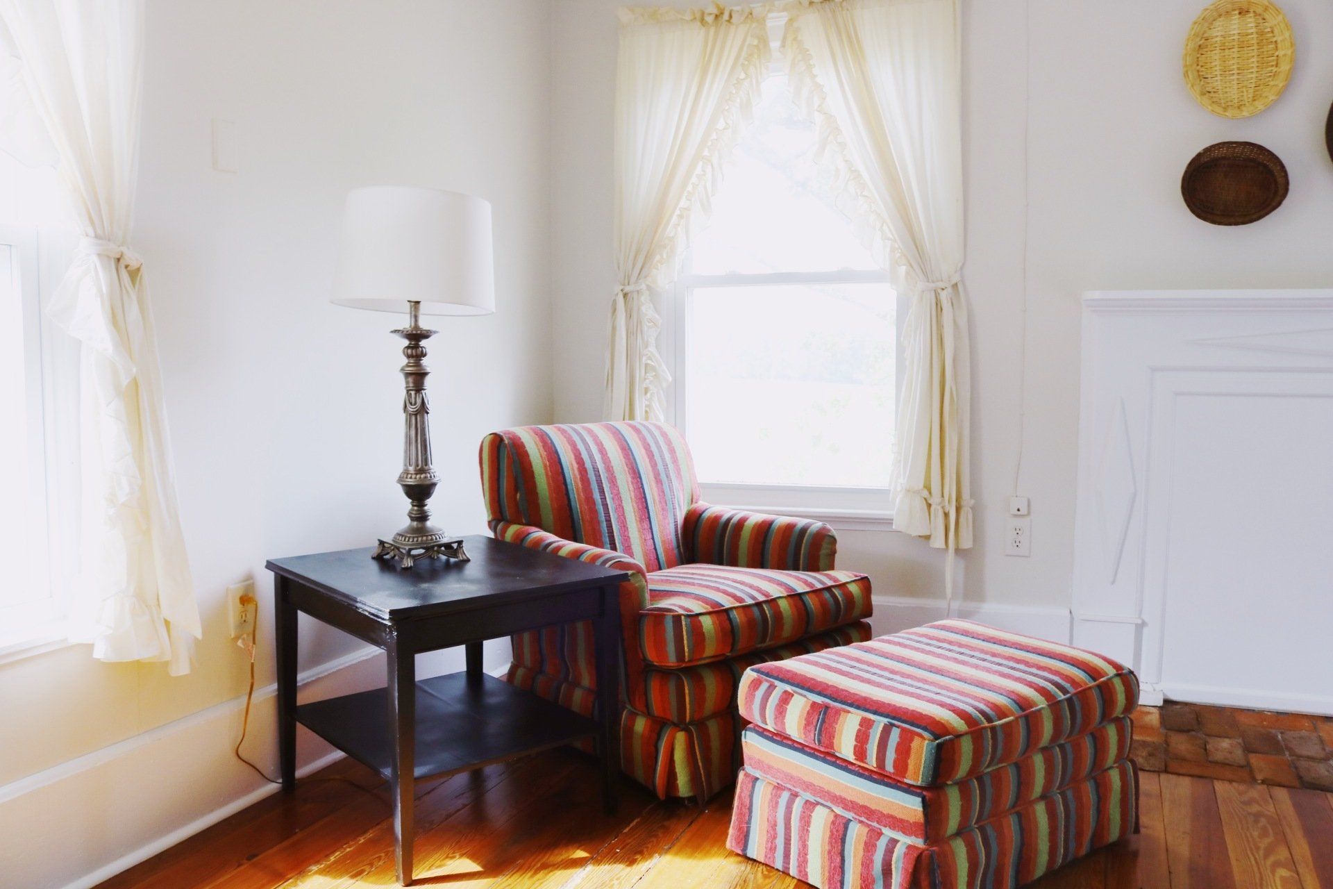 Cozy living area with striped armchair and ottoman, a table lamp, and window with curtains.