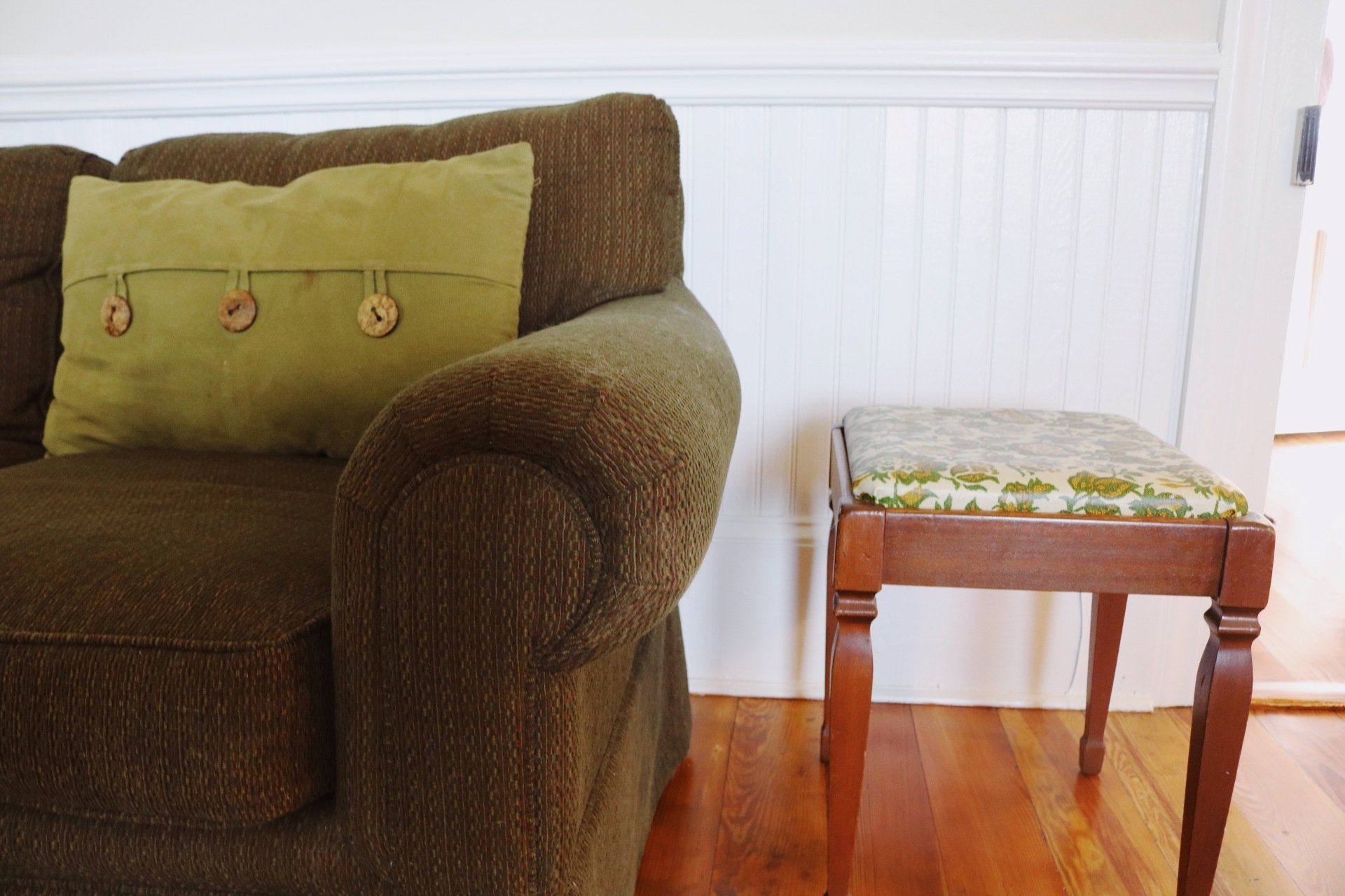 Brown couch with green pillow, next to a wooden stool, on a wood floor, near a white wall.