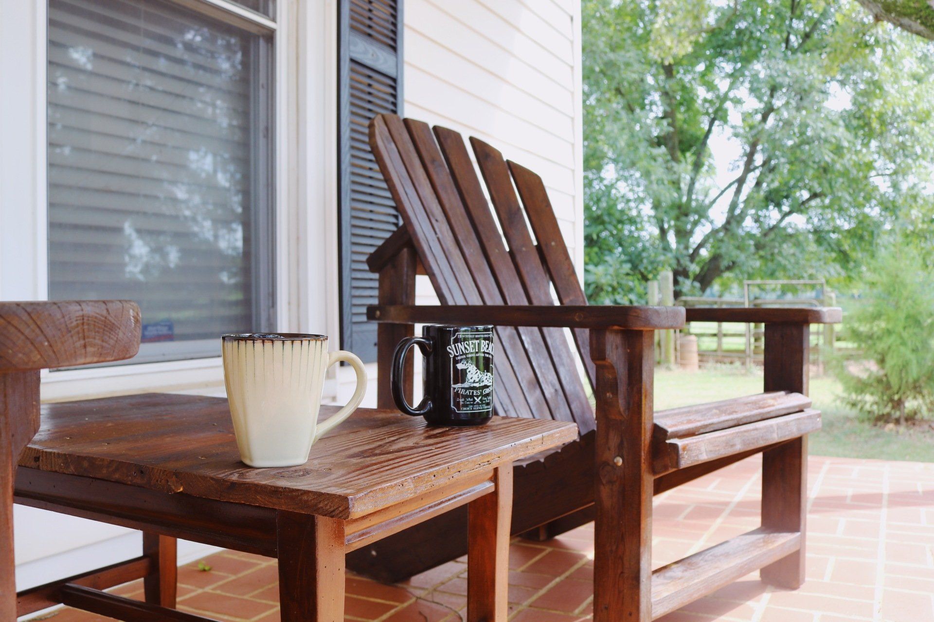 Wooden Adirondack chair and small table on a porch, with two mugs and a blurred outdoor background.