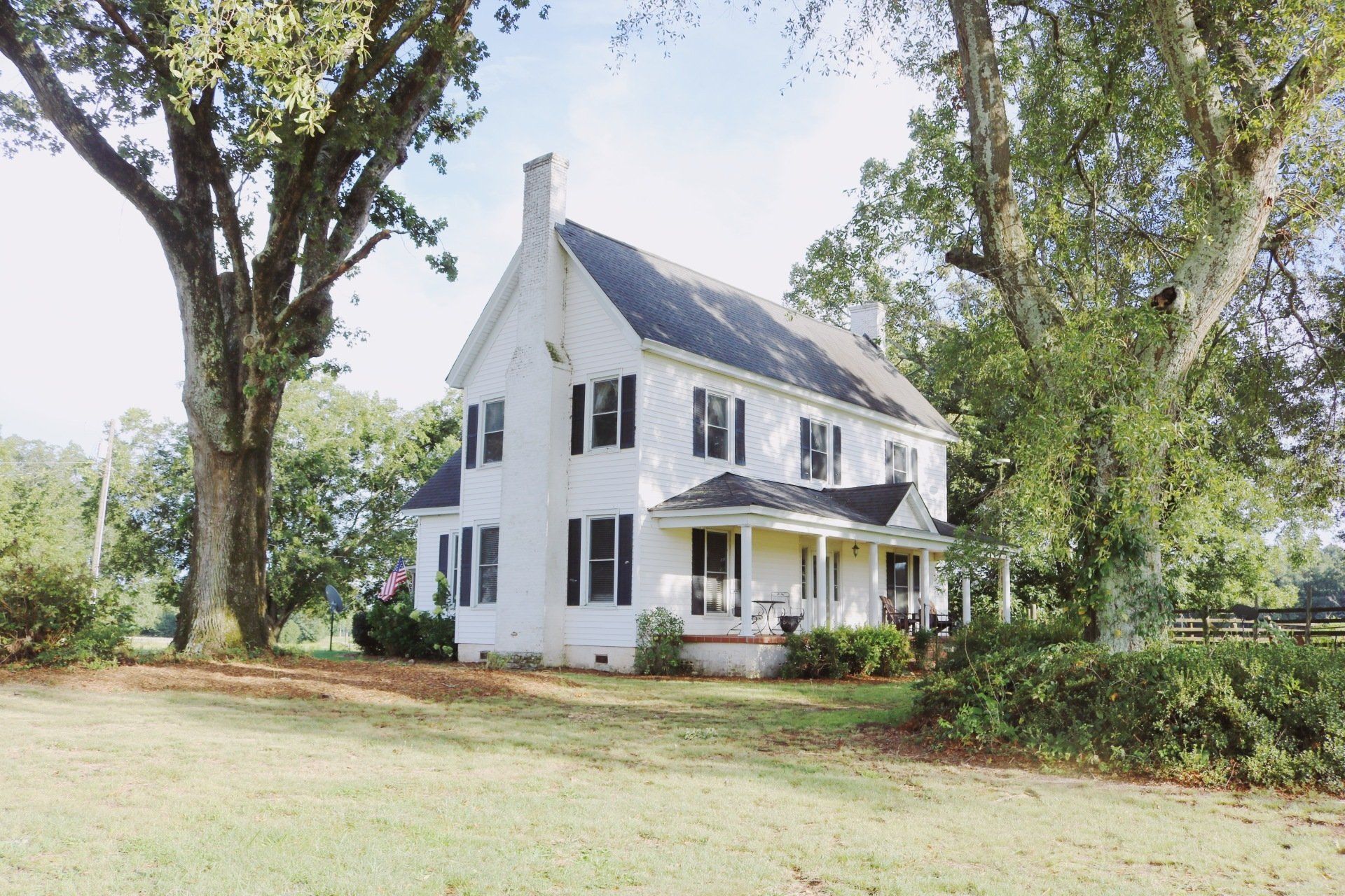 White two-story house with black shutters, tall trees on either side, grassy yard, and blue sky.