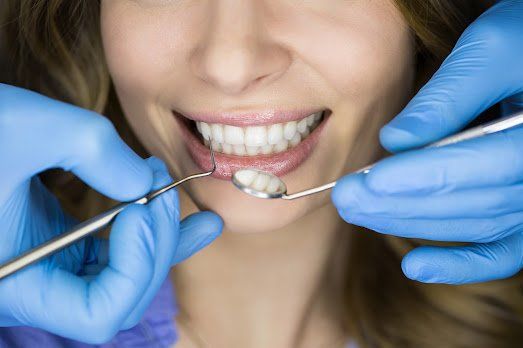 Dentist examining a woman's teeth with tools and blue gloves, patient smiling, close-up.