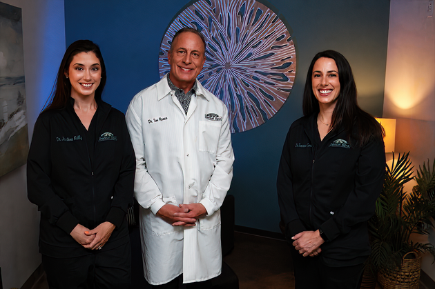 Four people in a dental office pose for a photo; three in black scrubs, one in a lab coat.