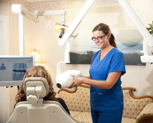 Dentist in blue scrubs smiling, using dental x-ray equipment on a seated patient in a dental office.
