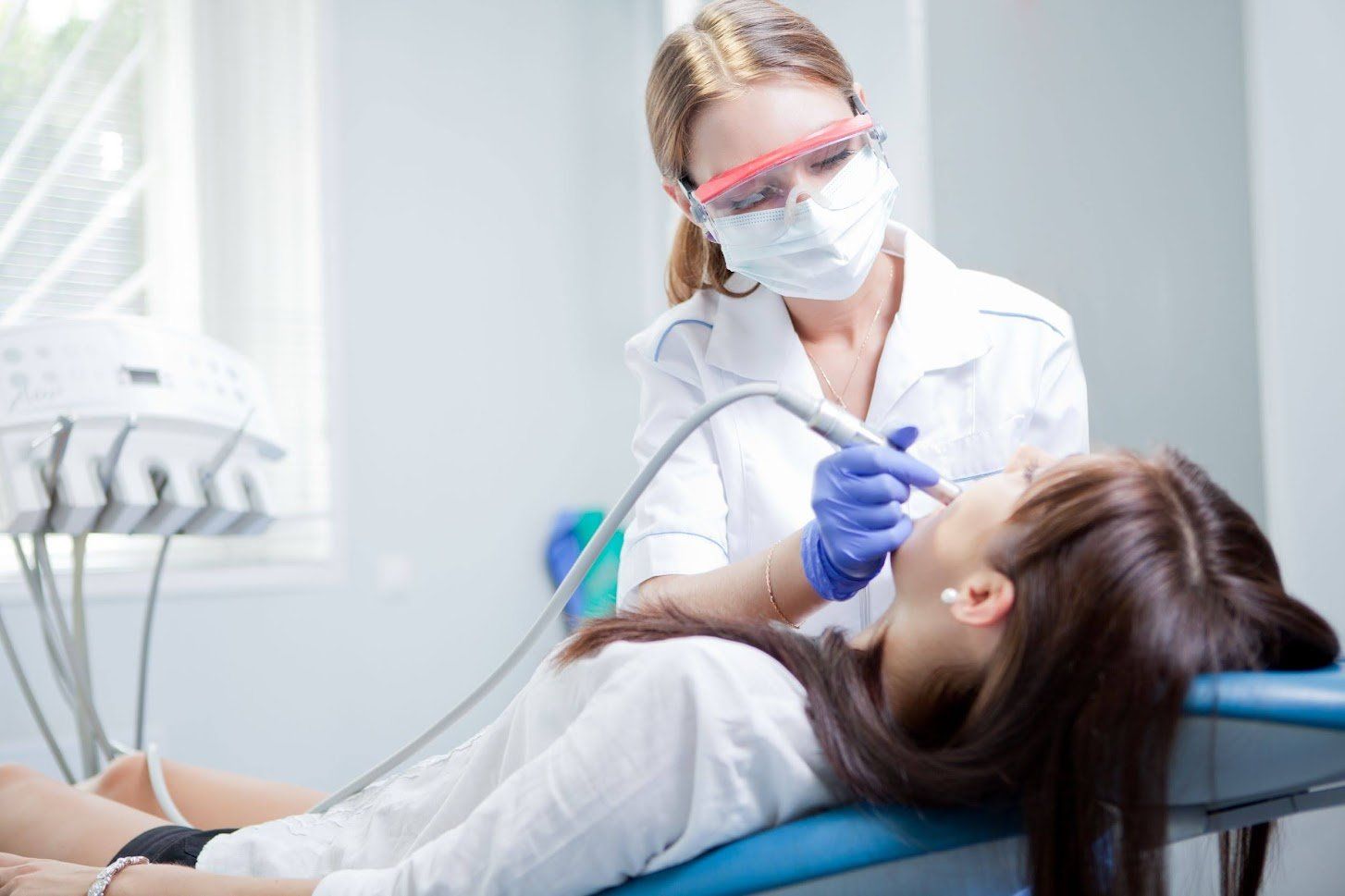 Dentist in mask and glasses examining patient's teeth in a dental office.