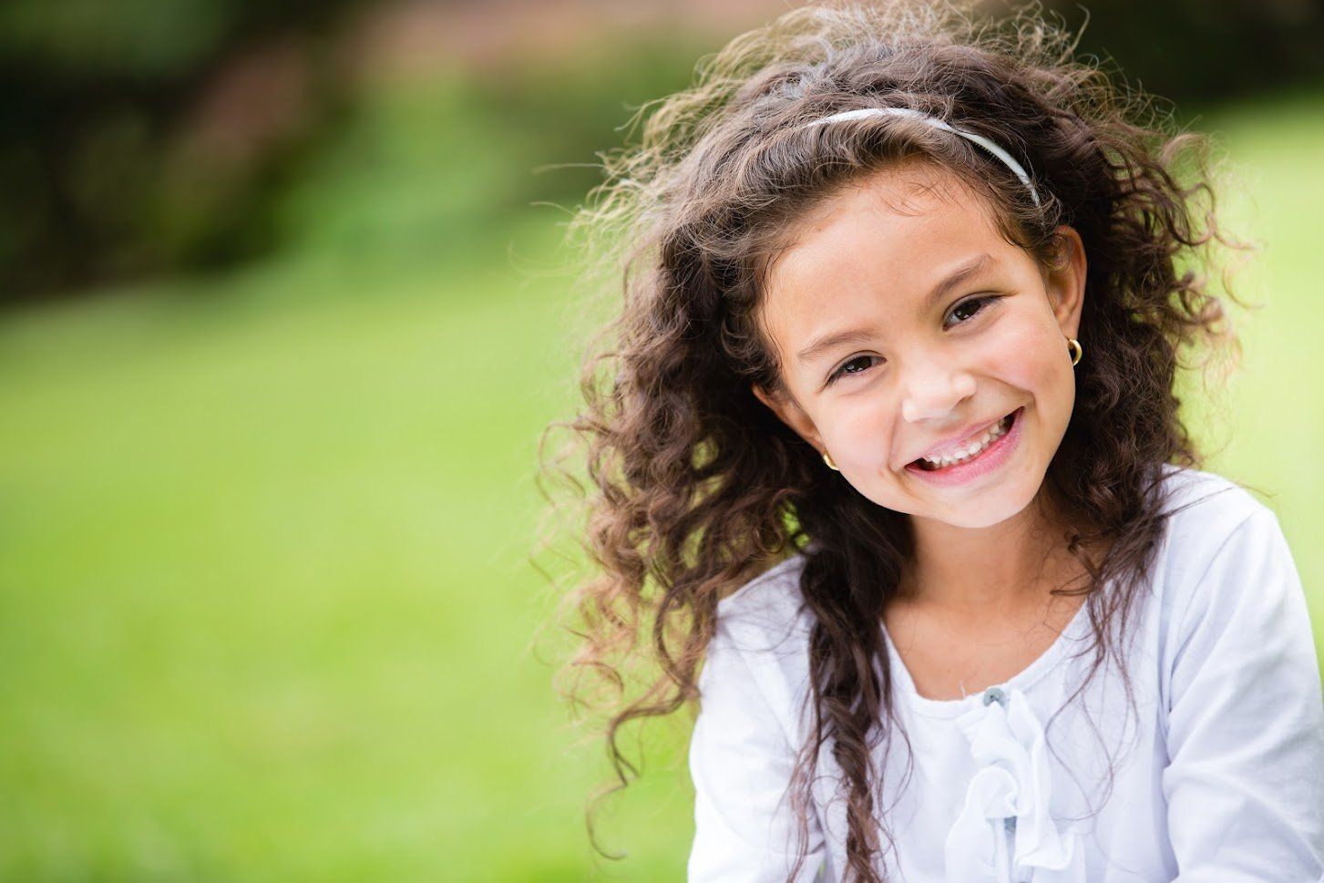 Smiling young girl with curly brown hair, wearing a white shirt and headband, outdoors.