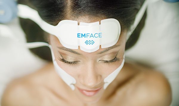 Woman undergoing EMface treatment; devices on forehead and cheeks, eyes closed.