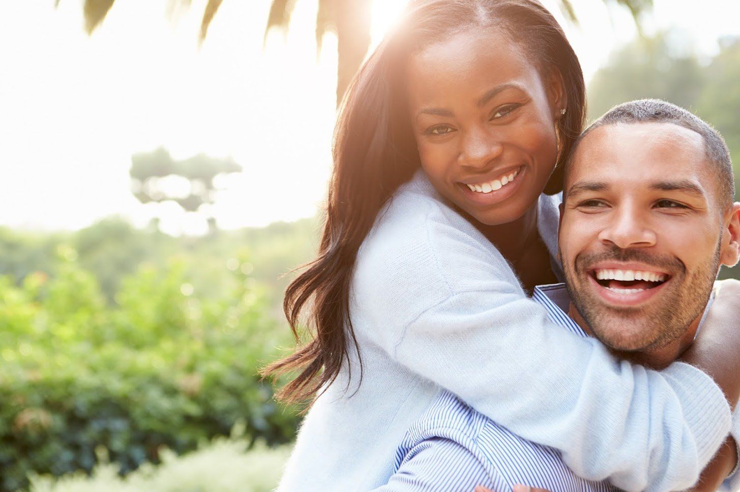 Smiling couple, woman piggybacking on man outdoors, backlit by sunlight.