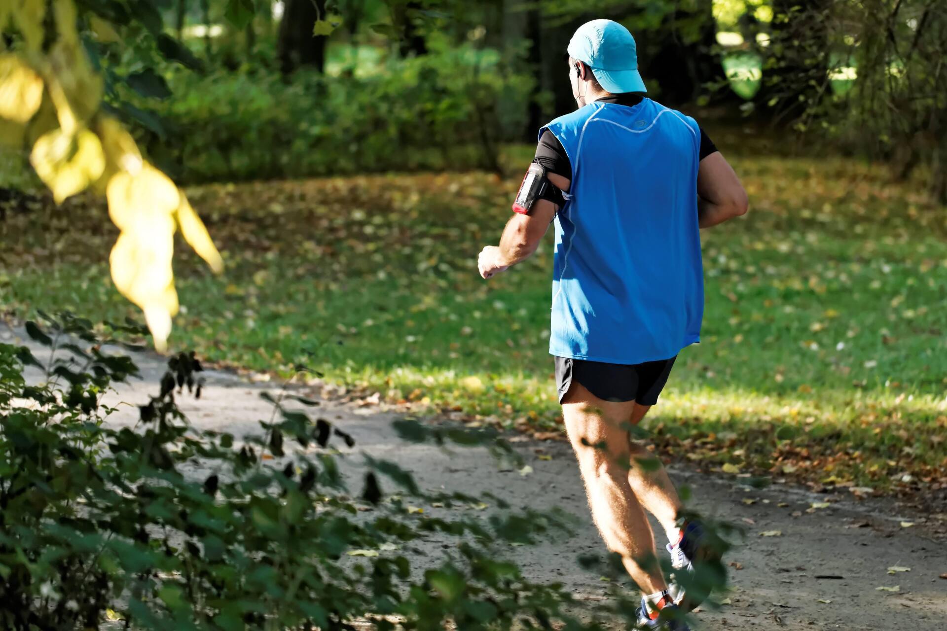 Man jogging on a path in a park, wearing blue hat and vest.