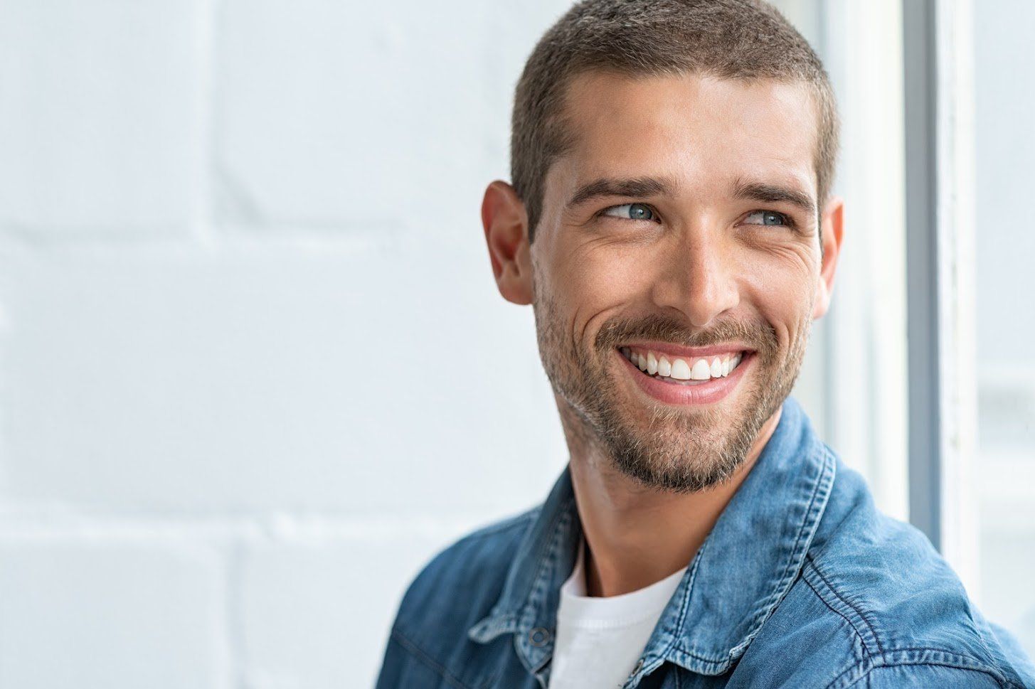 Man with a short haircut and beard smiles, wearing a denim shirt in a bright room.