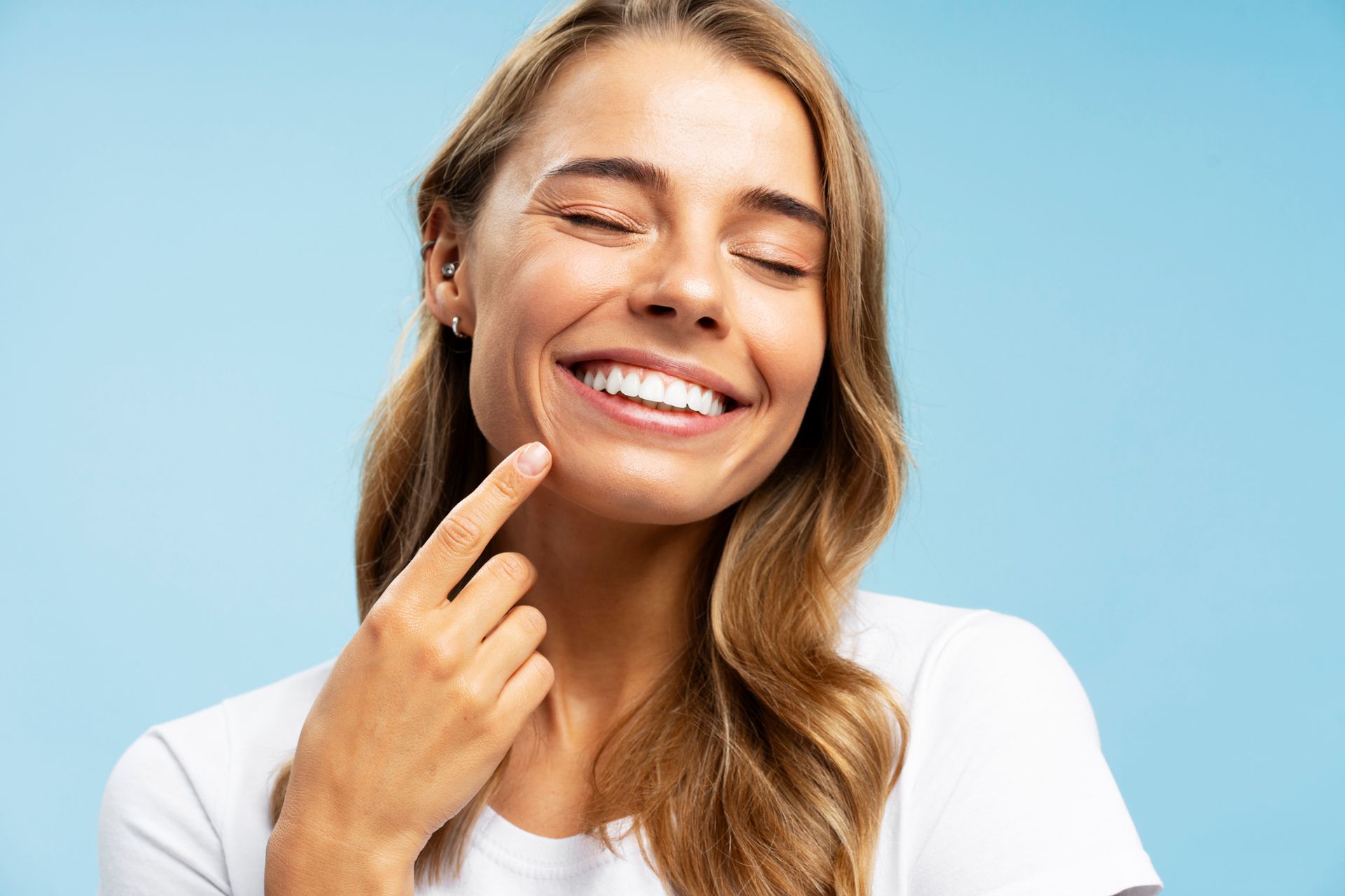 Person smiling with eyes closed, pointing to teeth against blue background.