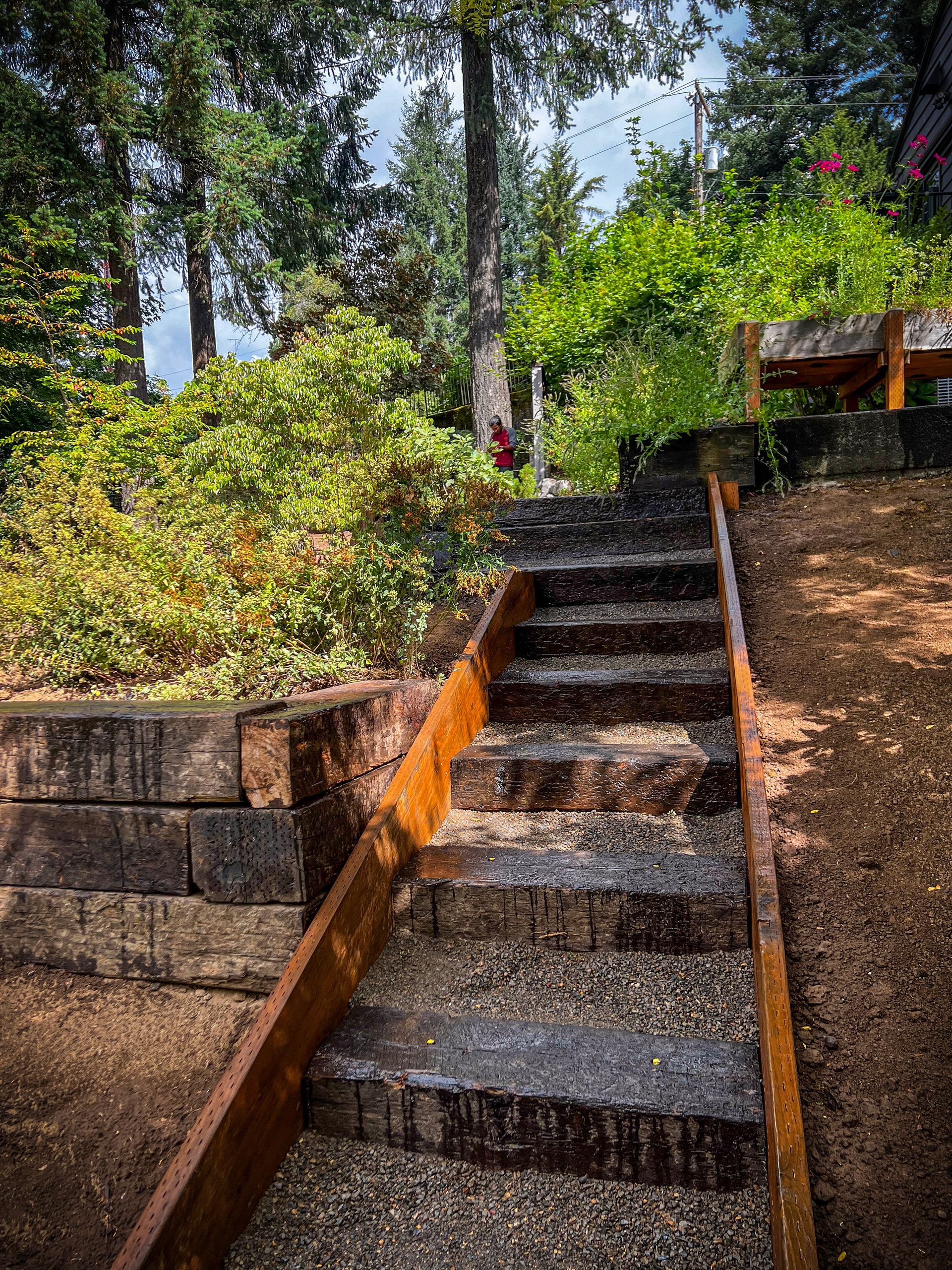 Stone steps with wooden handrail lead upwards in a garden, flanked by retaining wall and greenery.