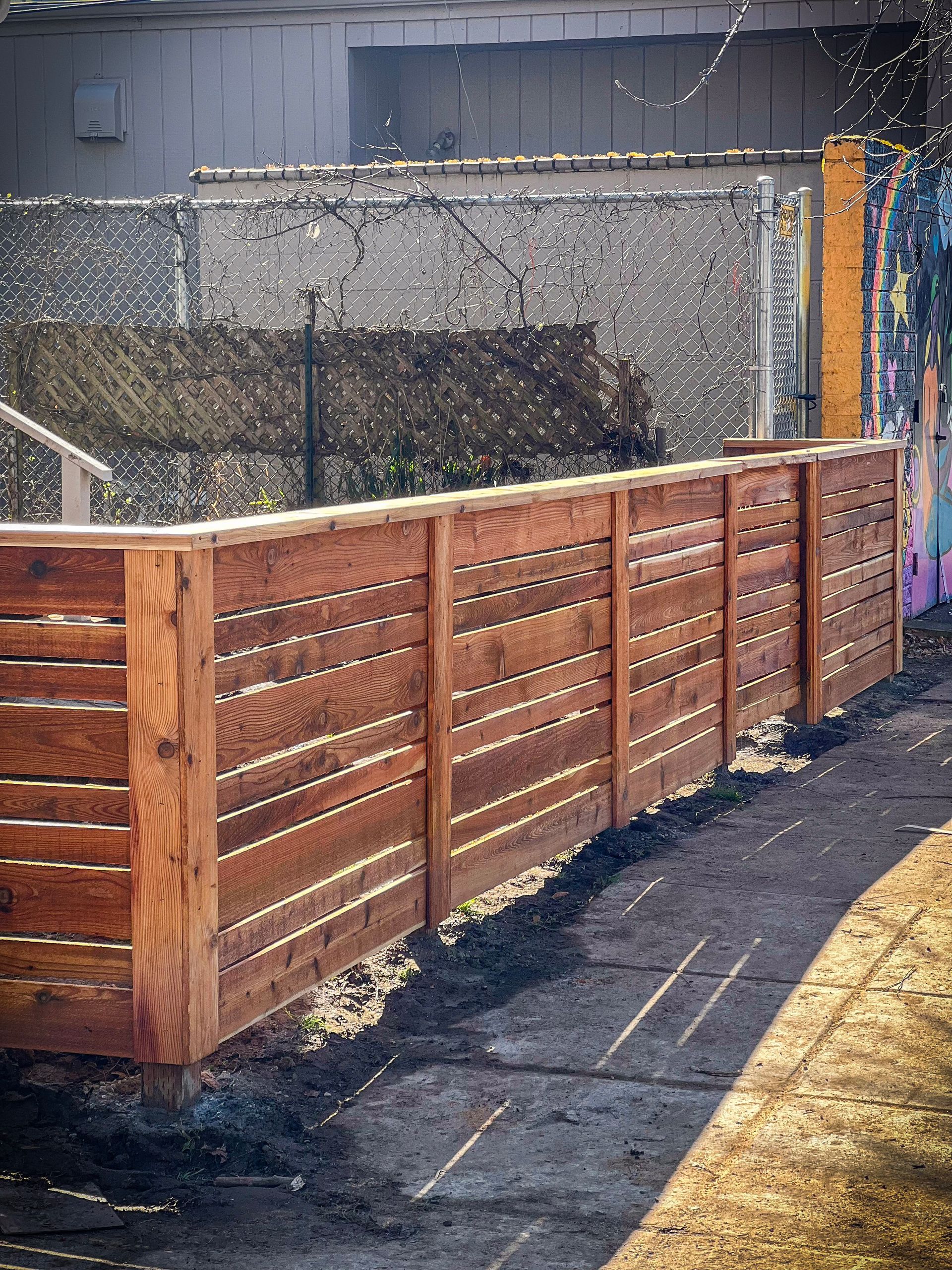 Wooden horizontal slat fence. The fence is brown and runs along a path next to a chain link fence.