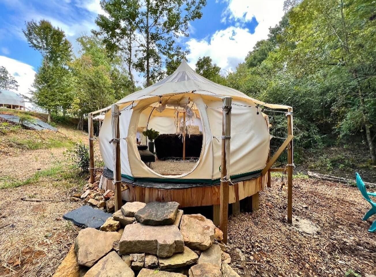 Beige glamping tent on a wooden platform surrounded by rocks, trees, and blue skies.