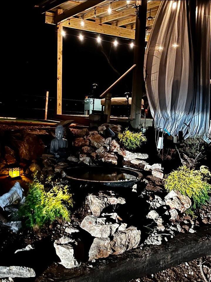 Water feature with rocks and plants under a wooden structure with string lights.