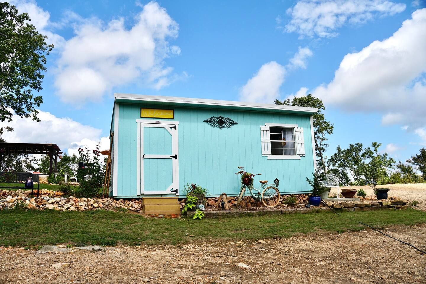 Turquoise shed with white trim, door, and window; blue sky and plants. A bicycle leans against it.